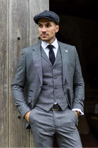 Man in a gray suit and cap standing against a wooden wall.